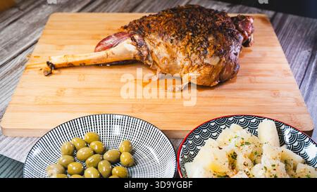 Gebratene Lammkeule auf einem Brett mit Beilagen: Oliven und Maniok-Pommes. Golden, gewürzt, perfekt für ein komplettes und herzhaftes festliches Essen. Stockfoto