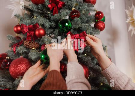 Großaufnahme von zwei Personen, die den Weihnachtsbaum mit roten Bögen und grünen Kugeln in einem gemütlichen festlichen Innenraum dekorieren. Stockfoto