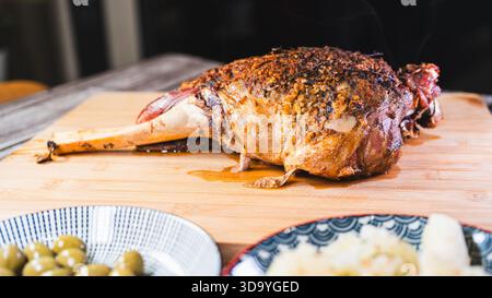 Gebratene Lammkeule auf einem Brett mit Beilagen: Oliven und Maniok-Pommes. Golden, gewürzt, perfekt für ein komplettes und herzhaftes festliches Essen. Stockfoto