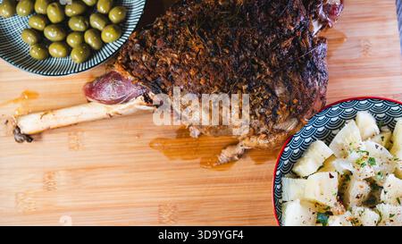 Gebratene Lammkeule auf einem Brett mit Beilagen: Oliven und Maniok-Pommes. Golden, gewürzt, perfekt für ein komplettes und herzhaftes festliches Essen. Stockfoto