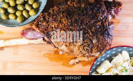 Gebratene Lammkeule auf einem Brett mit Beilagen: Oliven und Maniok-Pommes. Golden, gewürzt, perfekt für ein komplettes und herzhaftes festliches Essen. Stockfoto