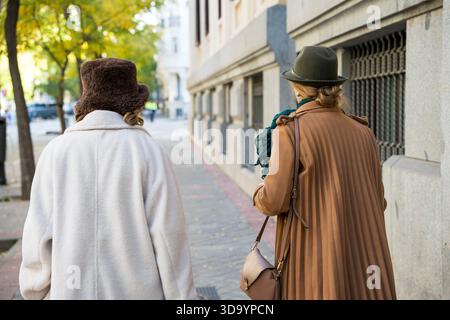 Zwei modische Frauen gehen Seite an Seite entlang einer von Bäumen gesäumten Straße, in kuscheliger Oberbekleidung gehüllt. Herbstblätter bilden eine farbenfrohe Kulisse und schaffen eine ruhige Atmosphäre Stockfoto
