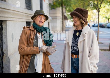 Zwei Frauen führen ein lebhaftes Gespräch, während sie durch eine geschäftige Straße schlendern. Sie tragen stilvolle Hüte und warme Mäntel und genießen das angenehme Wetter Stockfoto