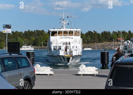 Nynashamn, Stockholmer County, Schweden - 29. Mai 2025: Eine kleine Fähre fährt durch einen Hafen mit mehreren Autos, die in der Nähe geparkt sind. Die Szene Stockfoto