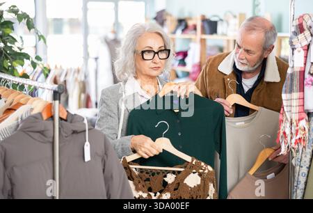 Ältere Ehepaare wählen im Bekleidungsgeschäft ein modisches warmes Kleid. Der Ehemann hilft seiner Frau, das Kleid zu wählen Stockfoto