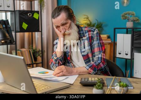 Müde erschöpfter Senior man Freelancer, der Papierkramme analysiert, Zahlungen abrechnet und monatliche Ausgaben für Bankrechnungen am Home Office Desk berechnet. Verärgerter Opa, der am Tisch arbeitet. Stockfoto