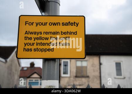 Ein gelbes Sicherheitsschild auf einem Bahnsteig, das die Fahrgäste anweist, hinter der gelben Linie zu bleiben, bis ihr Zug angehalten hat. Stockfoto