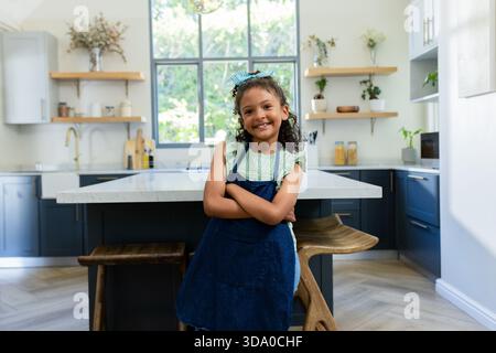 Hispanische Mädchen, die Arme kreuzten, lächelnd in der Küche mit Marmorinsel und Holzregalen Stockfoto