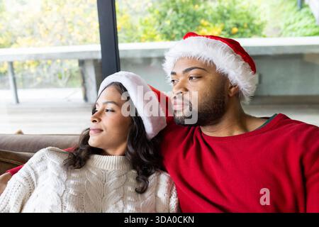 Verschiedene Paare sitzen auf einer gepolsterten Couch durch eine Glasschiebetür, tragen Santa-Mützen und Pullover Stockfoto