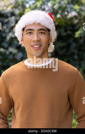 Asiatischer Mann an grüner Hecke mit rotem Weihnachtsmann-Hut, braunem gestepptem Pullover und weißem T-Shirt Stockfoto