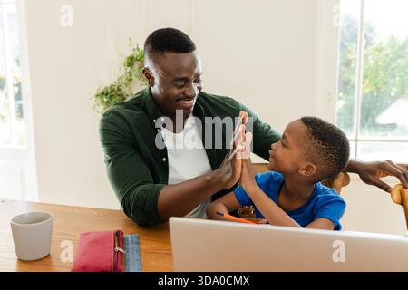 Afroamerikanischer Vater und Sohn sind High-Fiving am hölzernen Esstisch mit offenem Laptop Stockfoto