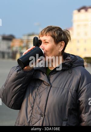 Die 60-jährige Russin trinkt Kaffee aus einem Thermalbecher im Freien in Wladiwostok und genießt warmes Sonnenlicht, ruhige Morgenstimmung und einfache achtsame Momente Stockfoto