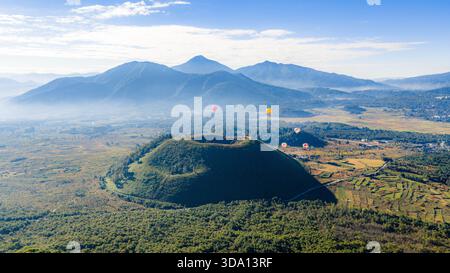 Vulkanischer Krater und vulkanischer Kegel im Tengchong Volcano Geopark mit Heißluftballons, die über das Basaltplateau in Yunnan, China, treiben. Stockfoto
