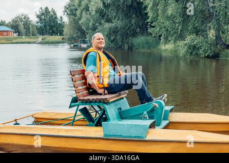Mann in oranger Schwimmweste, entspannt auf einem gelben Tretboot sitzend, das am Sommertag auf einem ruhigen Fluss schwimmt, umgeben von grünen Bäumen, Konzept der Outdoor-Re Stockfoto