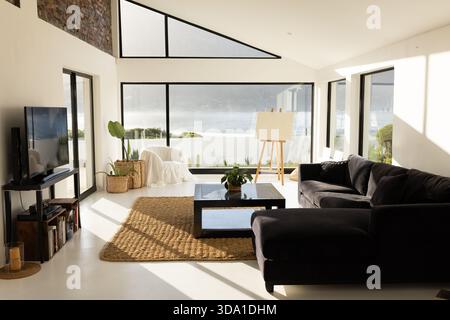 Dunkles L-förmiges Sofa mit Sonnenlicht am Wasser und schwarz gerahmten Fenstern Stockfoto