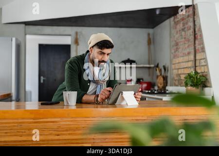 Indischer Mann mit Mütze, der sich an der Küchentheke beugt, mit Tablettenständer neben dem Becher, Smartphone Stockfoto