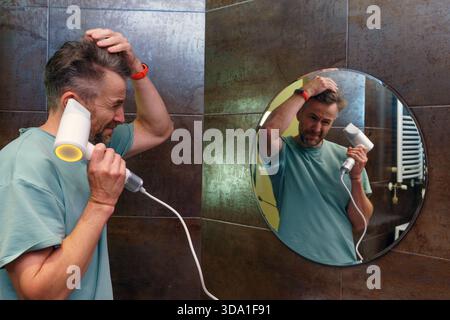 Ein Kaukasier mittleren Alters mit kurzen, grauen Haaren und einem Bart trocknet sein Haar mit einem weißen Haartrockner. Er wird im Profil gesehen, während seine Reflexion Stockfoto