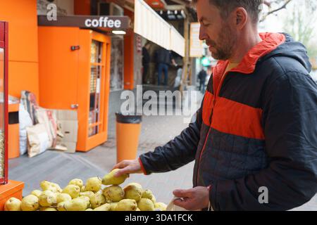 Ein Mann in einer schwarz-orangen Pufferjacke wählt eine frische gelbe Birne aus einem Obststand auf einem Markt in einer Stadtstraße. Stockfoto