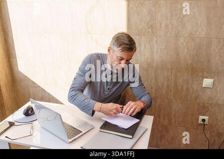 Ein fokussierter Mann mittleren Alters in einem grauen Hemd arbeitet an einem weißen Schreibtisch und schreibt mit einem Stift auf ein Dokument. Sein Heimbüro umfasst einen Laptop, ein Notebook, Stockfoto