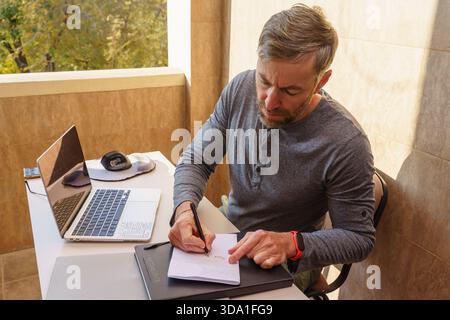 Ein konzentrierter Mann mittleren Alters sitzt an einem Schreibtisch auf einem sonnigen Balkon und schreibt auf einem Notizblock. Ein Laptop, eine ergonomische Maus und ein Smartphone befinden sich auf dem weißen Tisch. Stockfoto