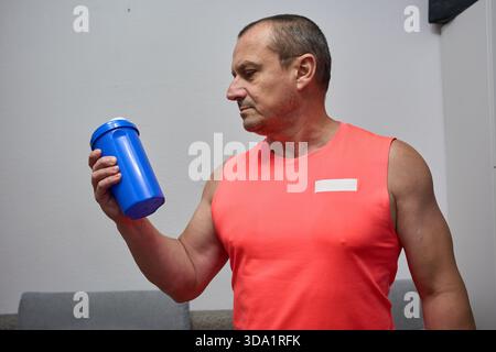 Mann mittleren Alters in rotem, athletischem Tanktop, der vor dem Workout eine blaue Protein-Shaker-Flasche hält Stockfoto