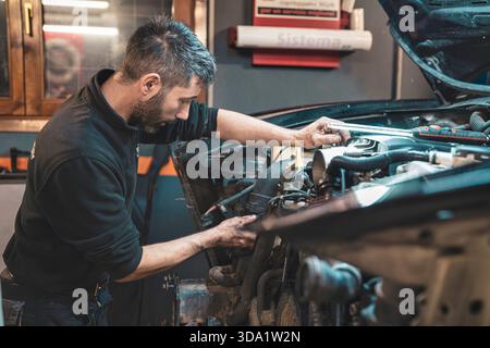 Rom, italien 20. november 2025: Professioneller Mechaniker, der in einer stark frequentierten Werkstatt einen Automotor inspiziert und repariert, mit Schwerpunkt auf Wartung und Diagno Stockfoto