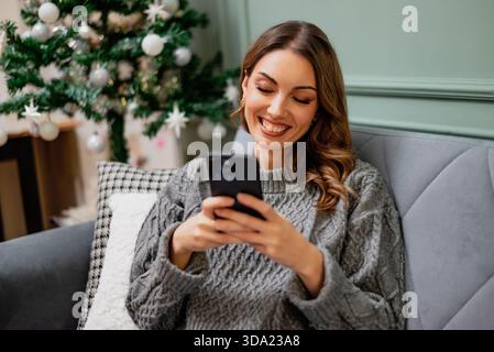 Die junge, glückliche lächelnde Frau sitzt auf der Couch in ihrem Haus und benutzt ihr Smartphone. Geschmückter Weihnachtsbaum im Hintergrund. Stockfoto