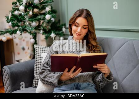 Eine entspannte Erwachsene Frau sitzt auf dem Sofa und liest ein Buch. Geschmückter Weihnachtsbaum im Hintergrund. Stockfoto