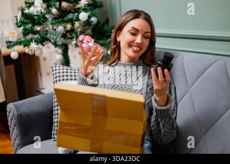 Glückliche Frau, die sich Parfüms ansieht, die sie online gekauft hat, und bekam ein Paket geliefert. Geschmückter Weihnachtsbaum im Hintergrund. Stockfoto