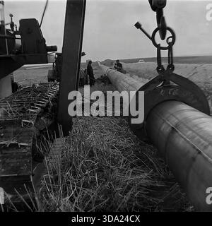 Region Donezk, Ukraine, UdSSR. Historisches Archivfoto aus den 1970er Jahren vom Bau der Gasleitung Schebelinka-Sloviansk. Eine Nahaufnahme mit niedrigem Winkel zeigt einen schweren sowjetischen Rohrverlegerhaken und eine Klammer, die ein Stahlrohr mit großem Durchmesser in einem Feld hält. Im Hintergrund schweißen die Arbeiter entlang der Grabenlinie Verbindungen. Diese Industrieszene verdeutlicht die enormen Größenordnungen und technischen Herausforderungen, die die Entwicklung der Energieinfrastruktur in der Donbass-Steppe während der Planwirtschaft der Sowjetzeit mit sich bringt. Stockfoto