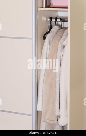 Weiche beige und weiße Wintermäntel hängen an Kleiderbügeln im modernen Kleiderschrank und symbolisieren die gemütliche saisonale Kleideraufbewahrung und das organisierte Zuhause Stockfoto