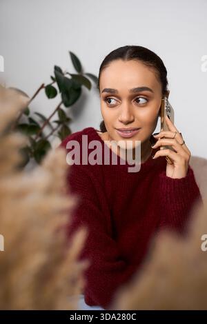 Eine junge Afroamerikanerin in einem kastanienbraunen Pullover plaudert am Telefon und fühlt sich wie zu Hause. Stockfoto