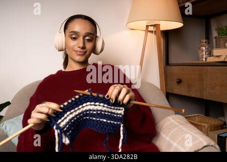 Eine junge Frau strickt gern in ihrem stilvollen Zuhause, während sie Musik hört. Stockfoto