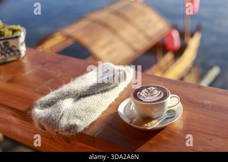 Gemütlicher, warmer Morgen mit einer Tasse Kaffee und Latte Art auf Holztisch. Ein Paar weiche Stricksocken neben dem Getränk sorgt für eine friedliche, entspannende Szene Stockfoto