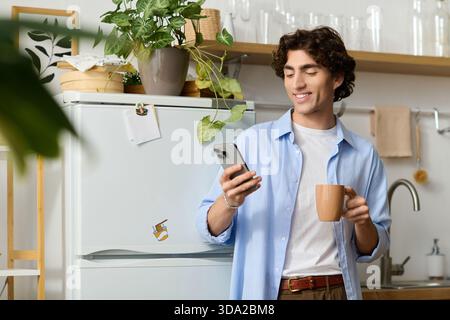 Der junge Mann lächelt, während er eine Kaffeetasse hält und drinnen sein Smartphone ansieht. Stockfoto