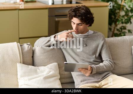 Ein hübscher junger Mann mit lockigen Haaren genießt seine Zeit zu Hause und konzentriert sich auf sein Tablet. Stockfoto