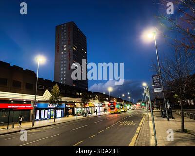 Salford, Großbritannien - 8. November 2025: Nächtliche Stadtstraße in Salford mit beleuchteten Geschäften, Bussen und einem hohen Turm. Stockfoto