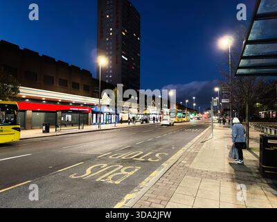 Salford, Großbritannien - 8. November 2025: Städtische Nachtszene in Salford mit Bussen, beleuchteten Ladenfronten und einem wartenden Fußgänger. Stockfoto