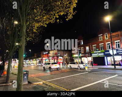 Salford, Großbritannien - 8. November 2025: Nächtliche Stadtstraße in Salford mit Autos, hellen Ladenschildern und Fußgängern. Stockfoto