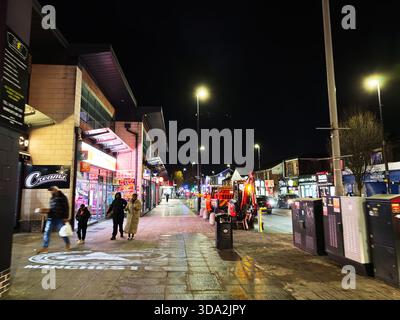 Salford, Großbritannien - 8. November 2025: Nachtszene im Stadtzentrum von Salford mit Shoppern, hellen Ladenfronten und einem Rettungsfahrzeug. Stockfoto