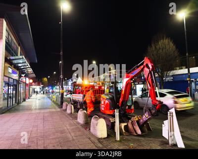 Salford, Großbritannien - 8. November 2025: Nächtliche Stadtstraße in Salford mit einem roten Bagger, Arbeitern in Orange und vorbeifahrenden Autos Stockfoto