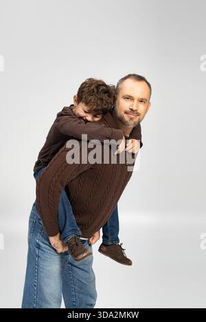 Ein Vater genießt einen verspielten Moment, indem er seinem Sohn einen Huckepack-Ritt mit einem Lächeln umgibt. Stockfoto