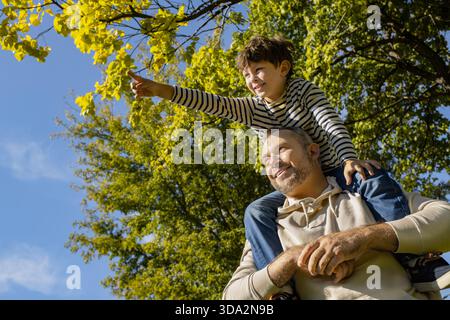 Ein Vater trägt seinen Sohn auf den Schultern, während er die Natur und das Lachen im Park genießt. Stockfoto