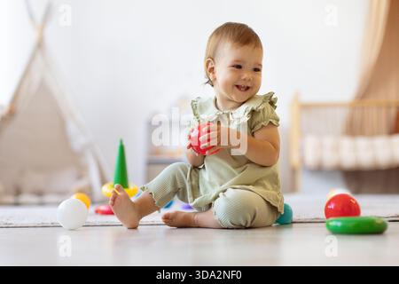 Baby hält roten Ball, während er auf dem Boden sitzt und von bunten Spielzeugen umgeben ist Stockfoto