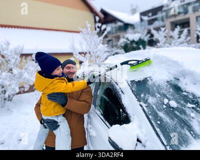 Ein Mann hält ein Kind in der Hand, während sie gemeinsam Schnee aus einem Auto entfernen, das in einem Wohngebiet geparkt ist. Schnee bedeckt den Boden und die umliegenden Häuser. Die s Stockfoto
