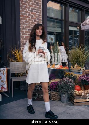 Modische junge Frau in weißem Strickkleid und schwarzen Plateauschuhen, die draußen laufen, eine Kaffeetasse halten, umgeben von einer lebhaften Herbsternte. Stockfoto