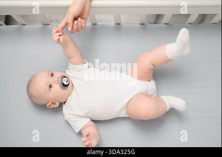 Baby mit Schnuller im Mund liegt auf dem Rücken im Kinderbett und hält die Hand von oben. Eltern spielen mit süßen Neugeborenen. Familie und Heim Konzept, Mutterschaft, Ma Stockfoto