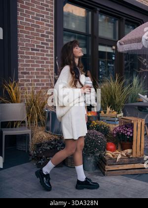 Modische junge Frau in weißem Strickkleid und schwarzen Plateauschuhen, die draußen laufen, eine Kaffeetasse halten, umgeben von einer lebhaften Herbsternte. Stockfoto