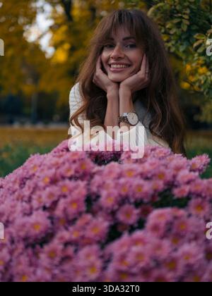 Nahaufnahme Porträt einer schönen jungen Frau, die fröhlich lacht und wegblickt, eingerahmt von hellrosa Chrysanthemen und gelbem Herbstlaub im Park. Stockfoto