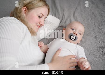 Frau Mutter liegt friedlich auf einem gemütlichen Bett mit einem kleinen Säugling, umarmt und streichelt ihn, Baby hat einen Schnuller im Mund. Mutterschaft, Säuglingsalter, Mutterschaft Stockfoto